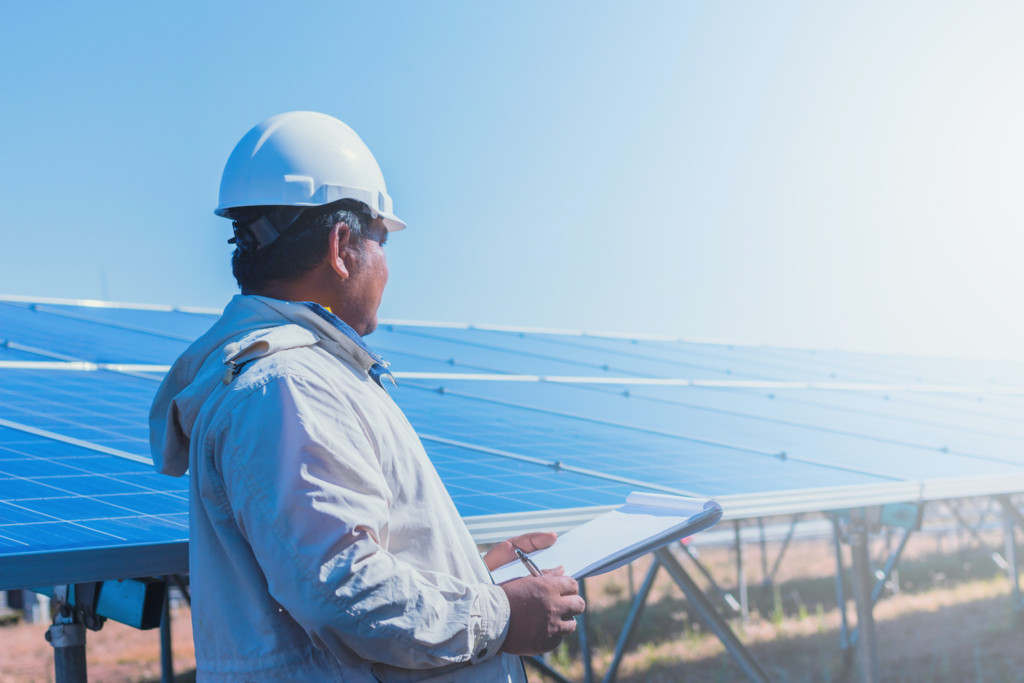 Man in hardhat holding clipboard looks at an array of solar panels